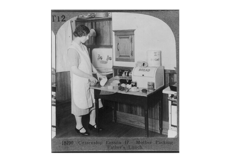 Stereo card depicting a woman in an apron and skirt preparing a meal. The original caption reads, "Citizenship Lessons: no. 17 - Mother Packing Father's Lunch."